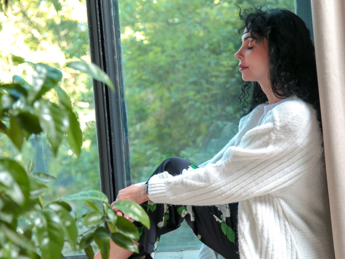 Una mujer con saco tejido blanco, sentada junto a la ventana de su casa con las rodillas recogidas y los ojos cerrados, en un momento de calma con luz natural y plantas alrededor.