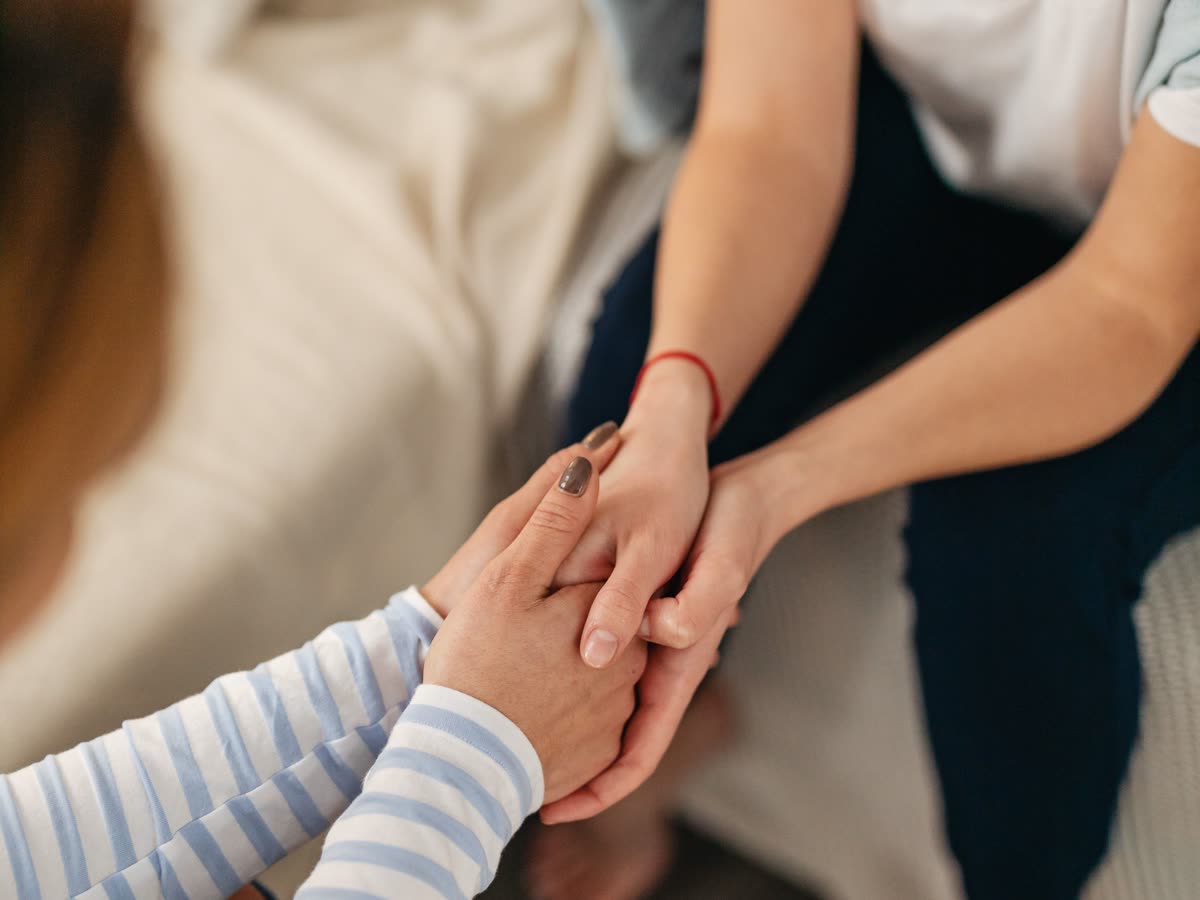 Dos mujeres sentadas una junto a la otra; una sostiene con ambas manos la mano de la otra, en un gesto tranquilo de acompañamiento, bajo luz natural suave.