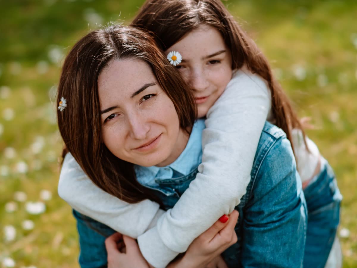 Una niña de unos once años abraza por detrás a su madre en un prado de margaritas; las dos miran a cámara con calma bajo la luz del sol.