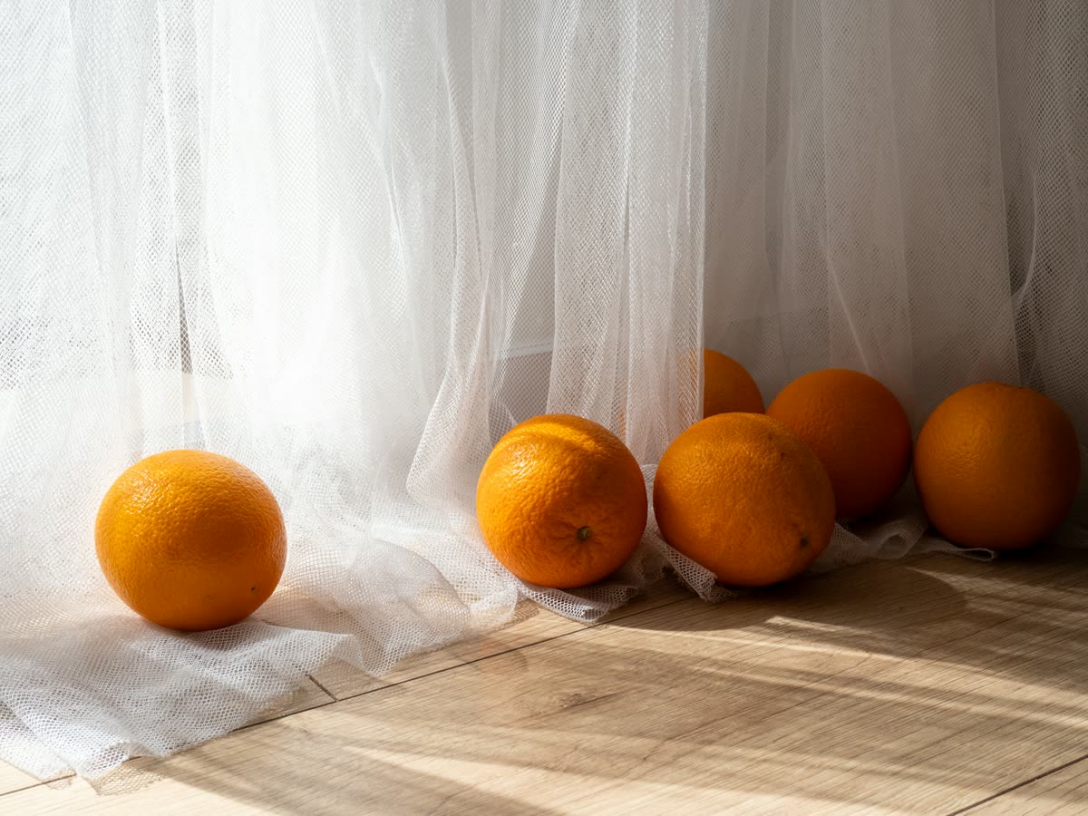 Varias naranjas enteras apoyadas en el suelo de madera clara junto a una cortina blanca translúcida, iluminadas por la luz suave de la mañana.
