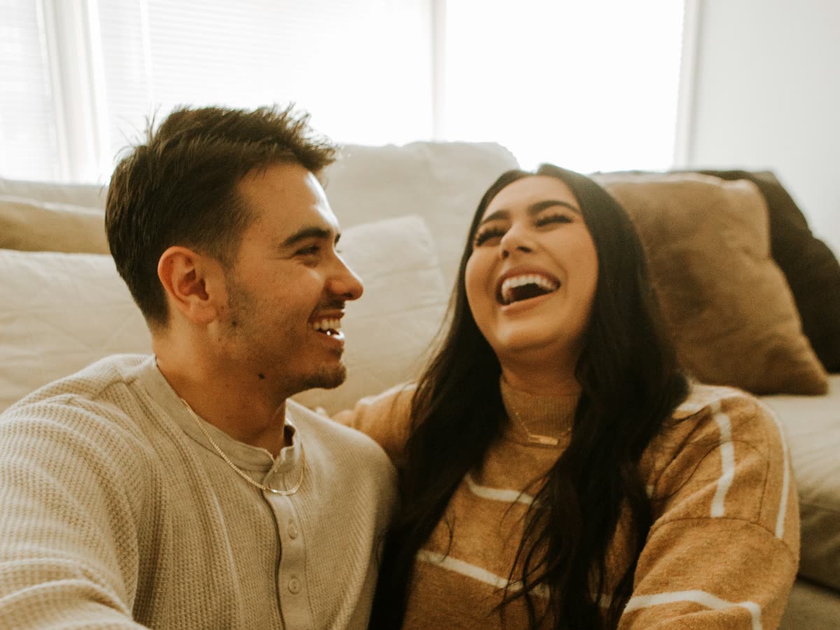 Una pareja joven ríe con complicidad, sentada en el suelo y apoyada en un sofá de una sala con luz natural.